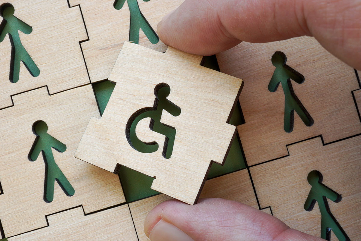 Mano coloca pieza de un puzzle de madera con el símbolo de accesibilidad en un tablero con figuras humanas.