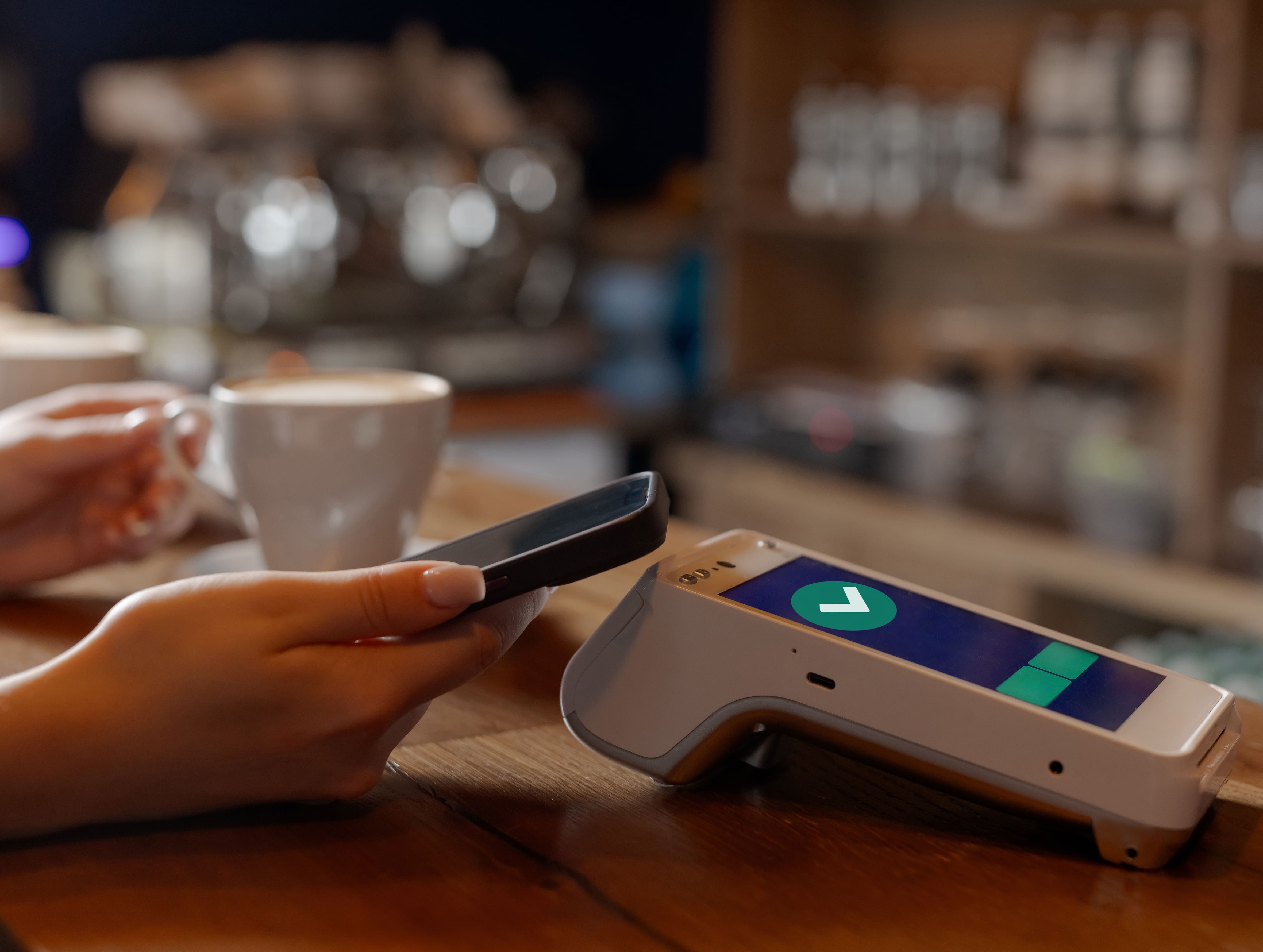 Mano de mujer acercando su móvil sobre un terminal para realizar un pago sobre una mesa con un café